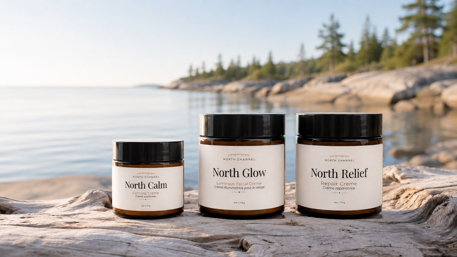 North Calm, North Glow, and North Relief crèmes standing on weathered driftwood, still water and a pine shoreline behind them